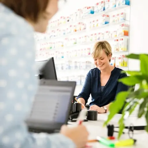 A smiling woman with short blonde hair interacts with a customer at a pharmacy counter. The background features organized shelves of medication. A second person is partially visible, working on a laptop, with a green plant on the table.