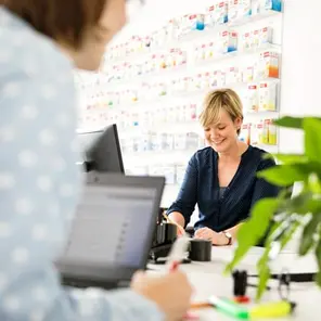 A smiling woman with short blonde hair interacts with a customer at a pharmacy counter. The background features organized shelves of medication. A second person is partially visible, working on a laptop, with a green plant on the table.