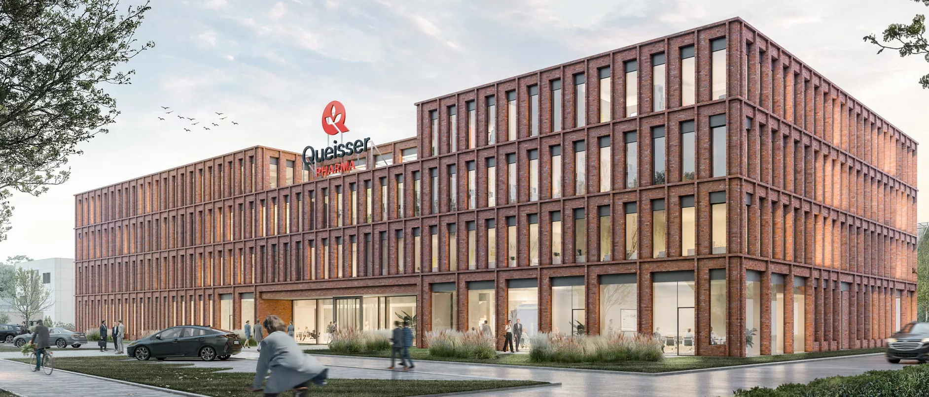 A modern, multi-story brick building features large windows and the sign "Queisser Pharma" prominently displayed on the roof. People are seen walking and cycling in front of the building, which is set amidst landscaping and vehicles parked along a paved pathway.