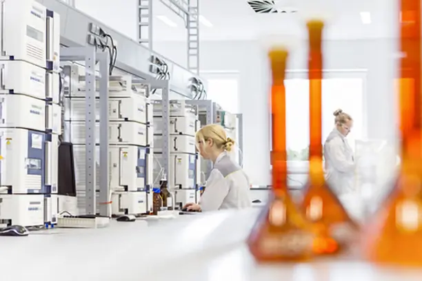 В лаборатории два ученых в белых халатах выполняют эксперименты. Один из них работает за компьютером, а другой занимается чем-то на заднем плане. Спереди видны колбы с оранжевой жидкостью. Лабораторное оборудование находится на полках.