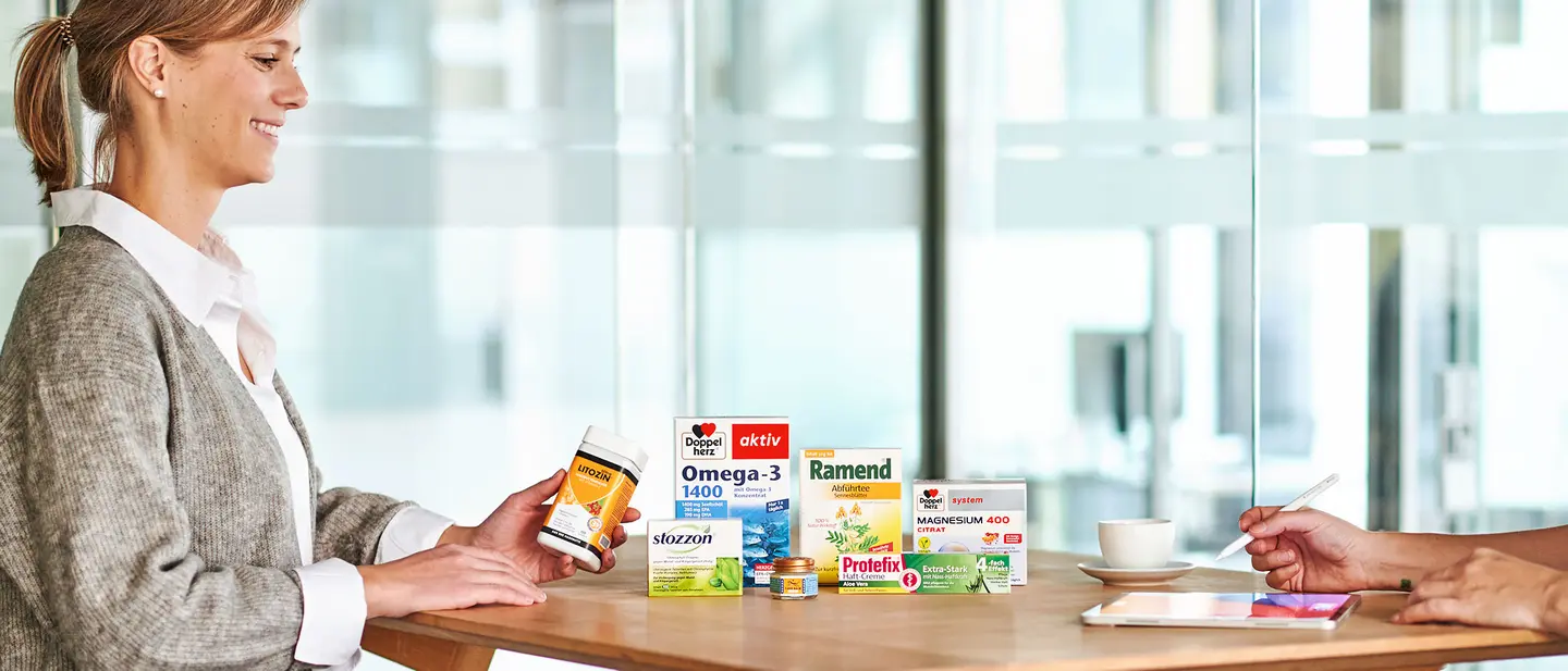 A woman smiles while holding a health supplement bottle at a table showcasing various product boxes. Another person is visible, writing on a tablet. A cup of coffee sits on the table, creating a collaborative atmosphere focused on health and wellness.