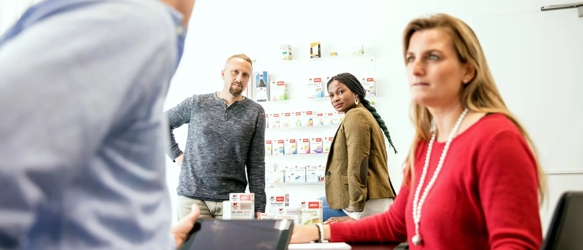 На изображении три человека в офисной обстановке. Две женщины и один мужчина обсуждают что-то, стоя рядом и сидя за столом. На заднем фоне видны полки с товарами. Все выглядят сосредоточенными и вовлеченными в разговор.